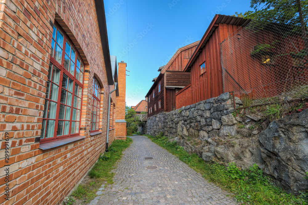 Narrow cobblestone street between well-preserved red brick building and traditional red wooden dwelling houses on stone hill. Stockholm, Sweden.