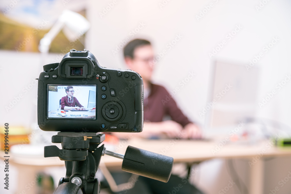 Video blogger at home: Camera on tripod is filming a young man sitting on his working place