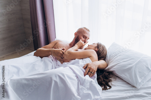 A loving couple laughs in bed. Smiling at each other on white sheets.