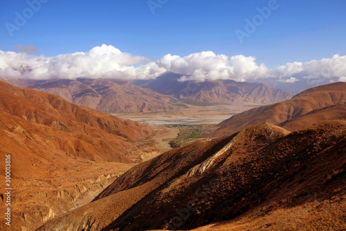 Panoramic view of the Tibetan Plateau showing brown mountains against a blue sky covered by white clouds.