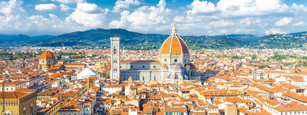 Fototapeta premium Górny panoramiczny widok z lotu ptaka na Florencję z katedrą Duomo Cattedrale di Santa Maria del Fiore, budynkami domów z pomarańczowo-czerwonymi dachówkami i pasmem wzgórz, błękitne niebo, białe chmury, Toskania, Włochy