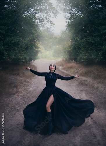 Gothic lady in black vintage silk flying dress, raises her hands in prayer. Beauty bare long leg. Witchcraft spell on the old road. Magic light and fog. Mysterious dark nature. Glamorous sensual Queen
