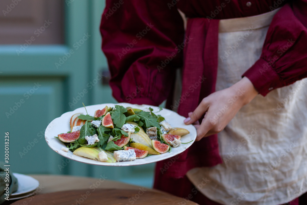 Young slim girl in red fress is holding white plate with delicious and healthy salad: green arugula, figs, pears, blue cheese. Tasty and low calories lunch. Bright colors, outdoors