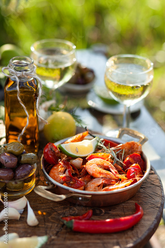 Romantic italian lunch outside for a couple: copper pan with delicious and spicy fried shrimps with herbs and garlic, wine, bread, olives. Bottle of olive oil. Luxury lifestyle, gourmet food