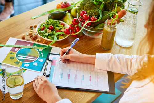 Woman dietitian in medical uniform with tape measure working on a diet plan sitting with different healthy food ingredients in the green office on background. Weight loss and right nutrition concept photo
