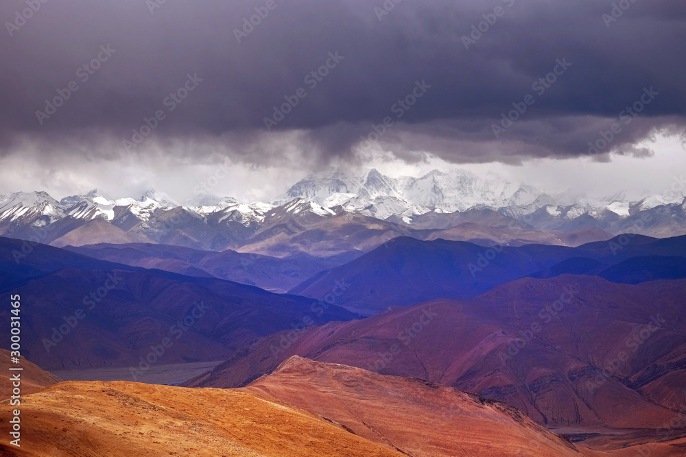 Panoramic view of the brown Himalaya Mountains, against the mountain range covered by snow and a dramatic sky.