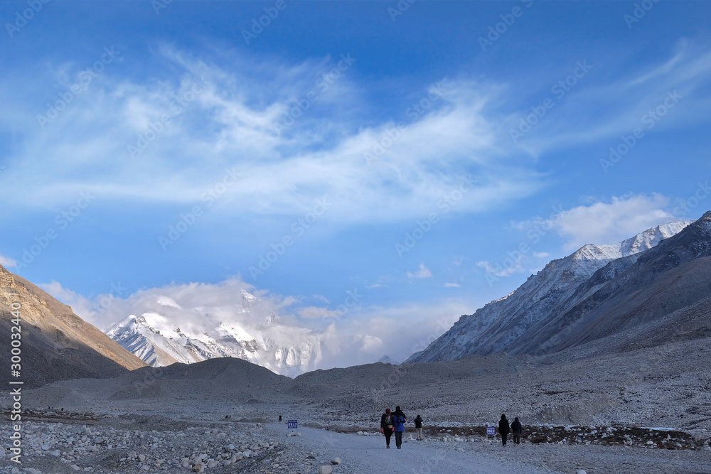 Tourists admiring Mount Everest at the Everest Base Camp in Tibet, against a blue sky covered by white clouds.