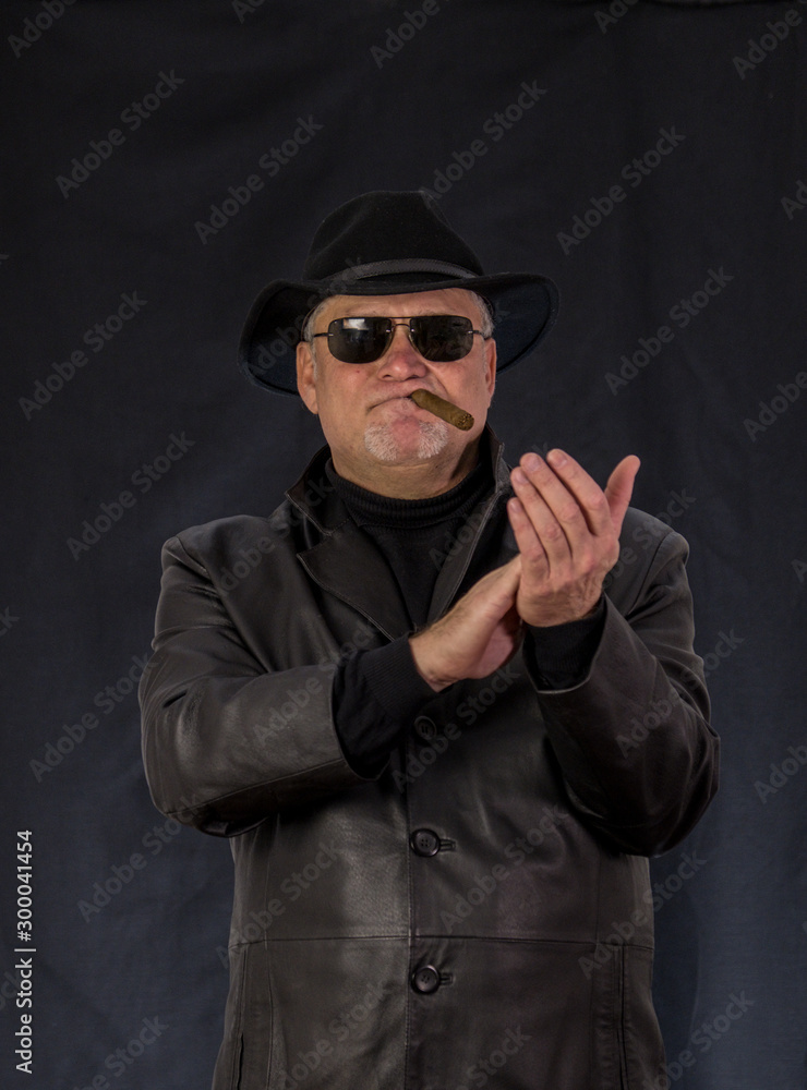 A man in black clothes, a black leather coat, hat. Ganster with a cigar. Mafiosi in black glasses on a dark background. Severe, strong, stylish, impressive, dangerous, unfriendly, evil, rude man. A man in black clothes, a black leather coat, hat. Ganster with a cigar. Mafiosi in black glasses on a dark background. Severe, strong, stylish, impressive, dangerous, unfriendly, evil, rude man.