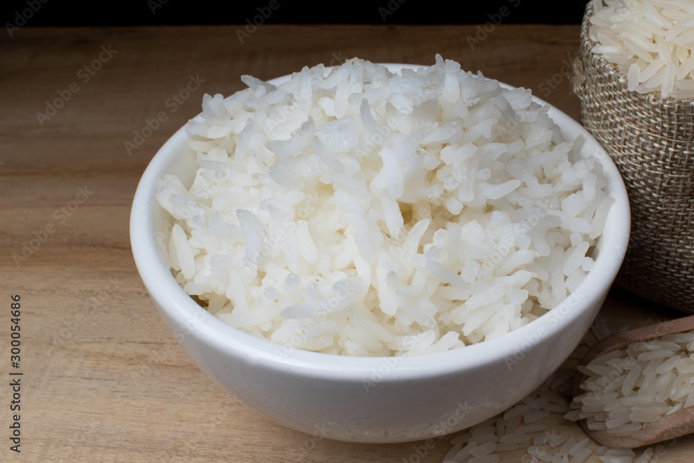 White ceramic cup with cooked rice Resting on a wooden table with a bag of rice grains and a wooden spoon, with a black backdrop. White ceramic cup with cooked rice Resting on a wooden table with a bag of rice grains and a wooden spoon, with a black backdrop.