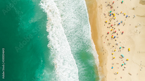 Aerial locked shot of waves breaking on the shore. Colourful beach umbrellas and people enjoying the summer.