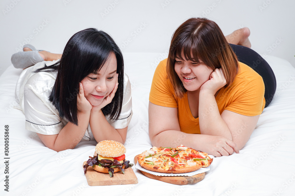 Two fat girls eat pizza and hamburgers in bed in the bedroom. Happy. The concept of eating and health care. Two fat girls eat pizza and hamburgers in bed in the bedroom. Happy. The concept of eating and health care.