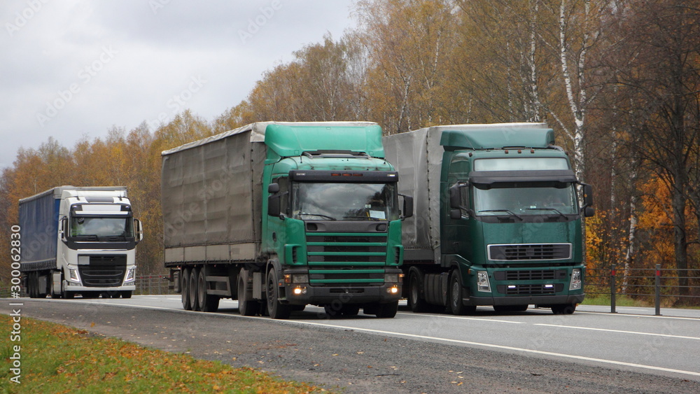 Transportation logistics, European green semi trucks with awning trailers move left to right on asphalt country road on autumn day, front side wide view