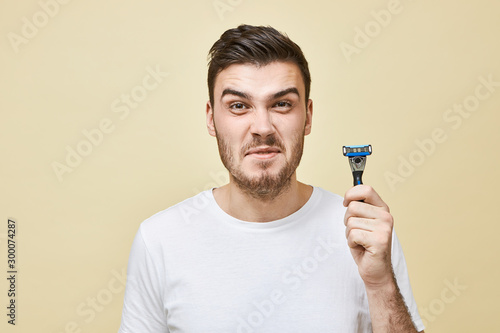 Studio image of unhappy displeased young brunette man with bristle grimacing does not want to shave his beard, hating shaving process, having sensitive skin, posing isolated with razor in hands