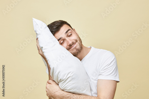 Horizontal image of handsome cute young man with bristle posing with head on white soft pillow sleeping peacefully and smiling, seeling good dream. Attractive guy napping after hard working day photo