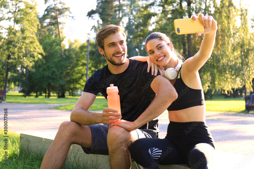 Young sporty couple in sportswear sitting on parapet in urban park and taking selfie after workout on summer sunny day. Young sporty couple in sportswear sitting on parapet in urban park and taking selfie after workout on summer sunny day.