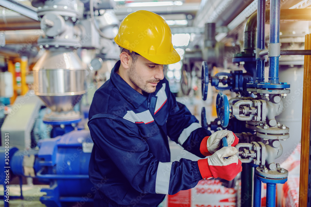 Dedicated hardworking worker in protective working clothes and with helmet on head screwing valve while standing in factory. Dedicated hardworking worker in protective working clothes and with helmet on head screwing valve while standing in factory.