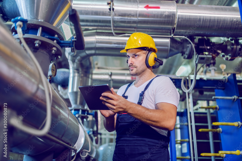 Serious handsome worker in overalls, with hardhat and antiphons using tablet while standing next to boiler in factory. Serious handsome worker in overalls, with hardhat and antiphons using tablet while standing next to boiler in factory.