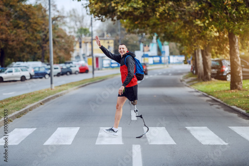 Full length of handsome caucasian handicapped sportsman in sportswear, with artificial leg and backpack crossing street and waving to a friend.