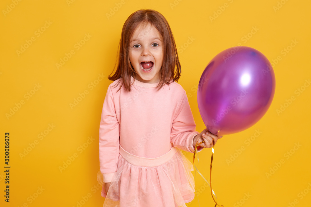 Portrait of surprised little girl with dark staright hair standing over yellow studio background beautiful clothes, holding purple ballon in hands and looking at camera joyfully. Holiday concept. Portrait of surprised little girl with dark staright hair standing over yellow studio background beautiful clothes, holding purple ballon in hands and looking at camera joyfully. Holiday concept.