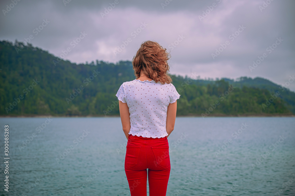 Young woman standing enjoying nature watching an amazing turquoise lake with mountains in the background on a cloudy day Young woman standing enjoying nature watching an amazing turquoise lake with mountains in the background on a cloudy day