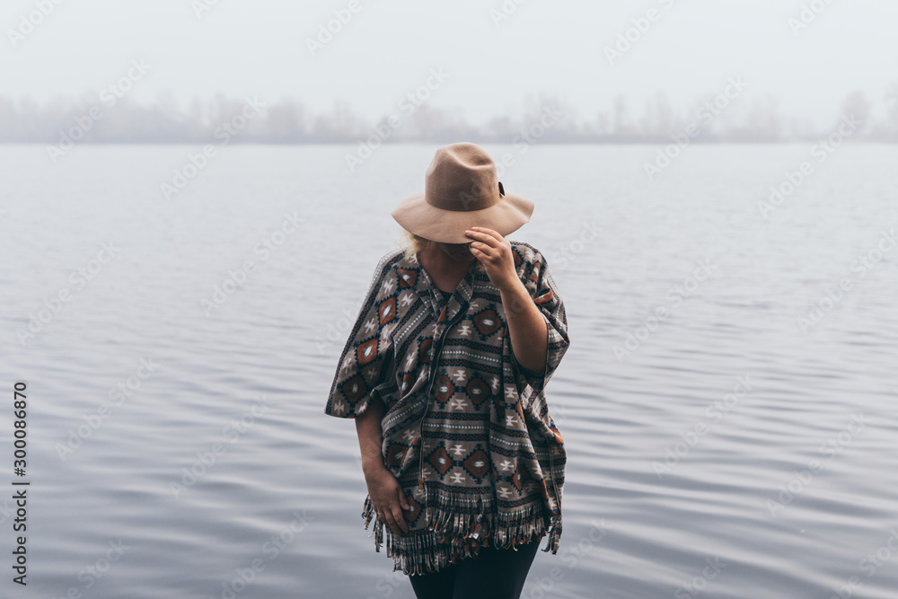 Obraz premium Woman in hat and poncho standing on the river bank and looking towards autumn forest covered with morning fog