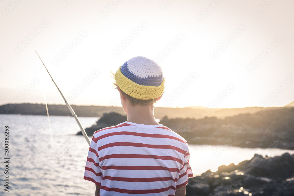 Back view of a young fisherman with with fishing pole in hand looking the sea. Sailor boy with wool cap and a striped t-shirt enjoying a summer evening. Lifestyle, outdoors, fun and carefree concept Back view of a young fisherman with with fishing pole in hand looking the sea. Sailor boy with wool cap and a striped t-shirt enjoying a summer evening. Lifestyle, outdoors, fun and carefree concept