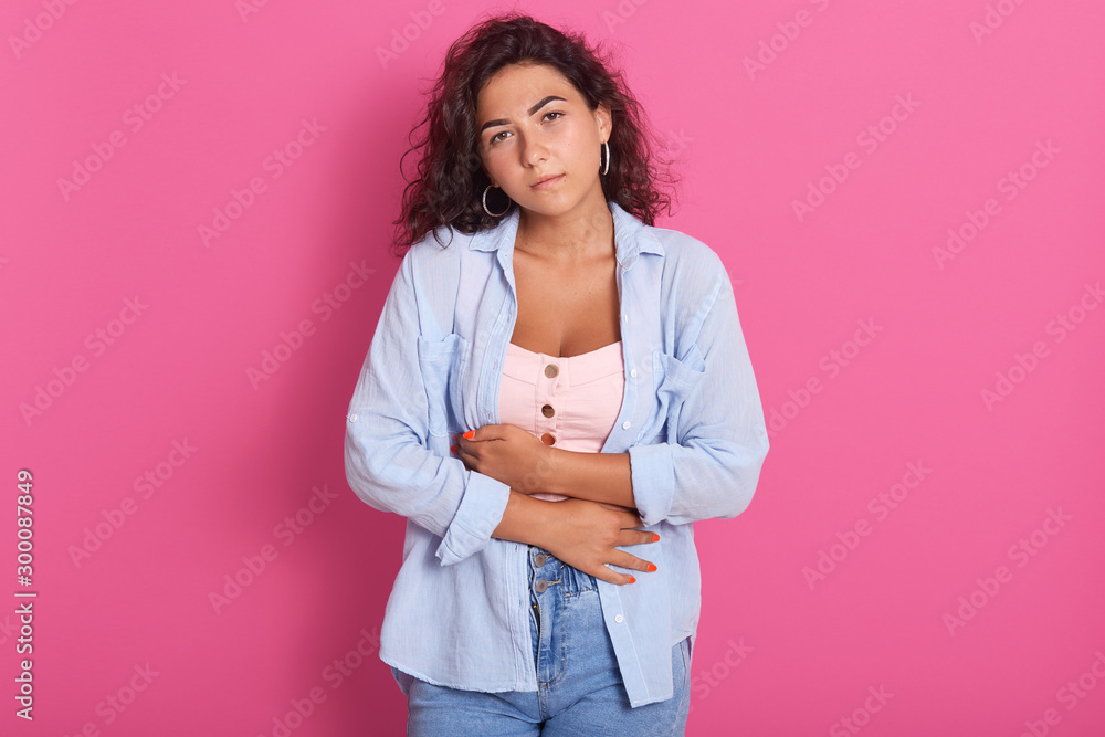 Close up portrait of pretty woman presses hands on belly, feels pain, having problems with health, wearing pink top, blue shirt and jerans, poses against pink background, grimaces from pain. Close up portrait of pretty woman presses hands on belly, feels pain, having problems with health, wearing pink top, blue shirt and jerans, poses against pink background, grimaces from pain.