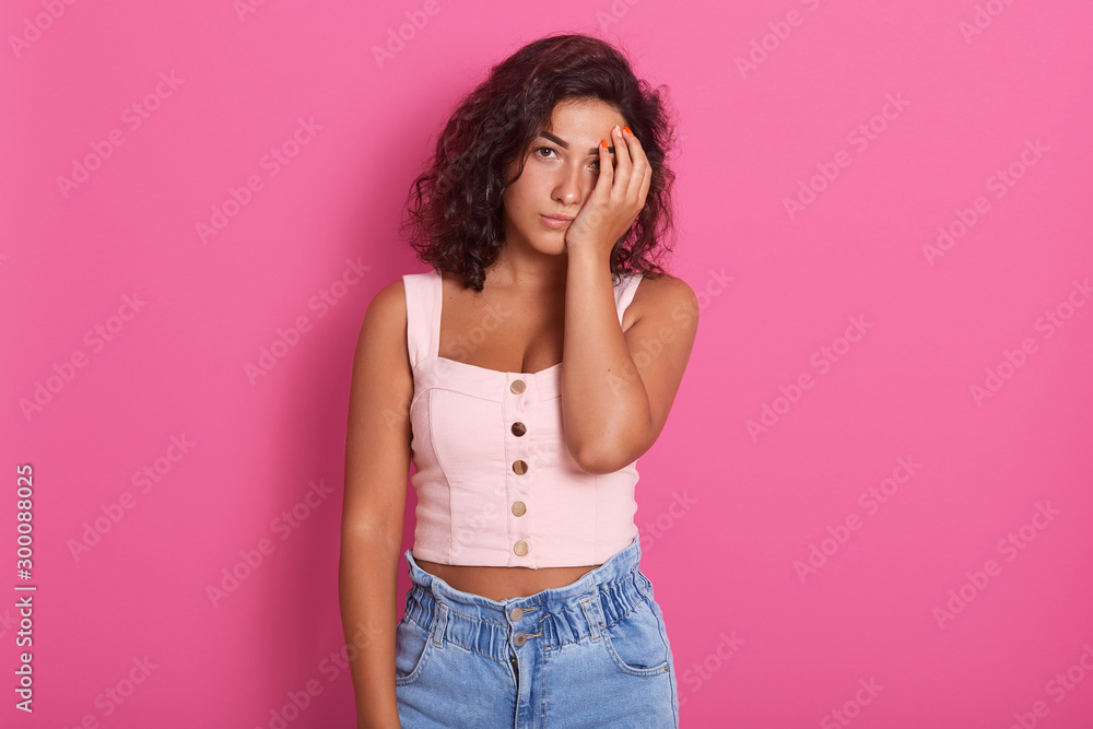 Horizontal shot of young winsome woman covers face with hand, has sleepy tired displeased facial expression, feels apathy, wearing stylish clothing, posing isolated over pink studio background. Horizontal shot of young winsome woman covers face with hand, has sleepy tired displeased facial expression, feels apathy, wearing stylish clothing, posing isolated over pink studio background.