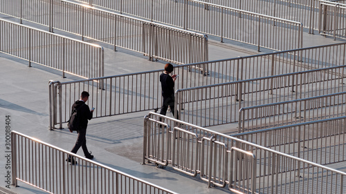 Rear view of two unrecognizable man and woman in black clothes walking and watching their mobile phones across metal hand railings used to separate queue lines.