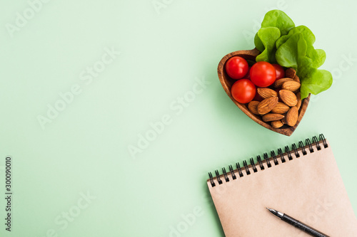 Concept of healthy eating, diet and new resolutions. Notebook and heart shaped plate with vegetables and nuts on a light green background. Top view, flat lay, copy space
