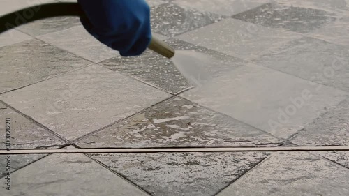 Hand of man wearing blue rubber gloves using a hose to cleaning the tile floor.