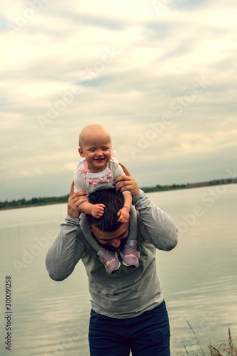 A child on his father’s neck. Walk near the water. Baby and dad against the sky.