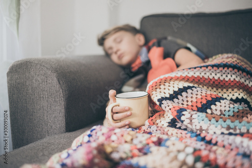 hot water bottle and a mug. Sad teen with the flu rests alone at home in a cold winter day. Child with seasonal infections and fever Soft focus on the cup., Sick boy sleeping with woolen blanket