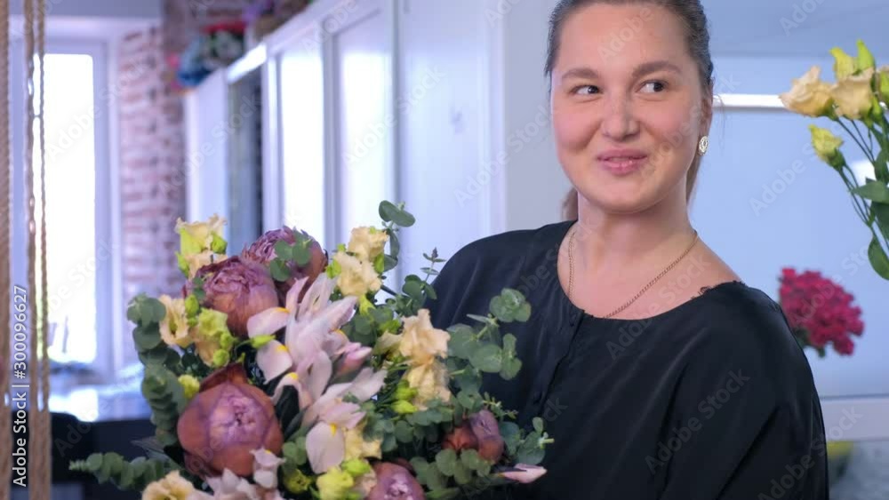 Floral business concept. Florist woman makes big flower bouquet of eucalyptus branches, beige eustoma, brown maroon peony and blue iris and smiles at camera in flower shop. Create professional bunch. Floral business concept. Florist woman makes big flower bouquet of eucalyptus branches, beige eustoma, brown maroon peony and blue iris and smiles at camera in flower shop. Create professional bunch.