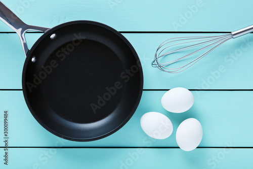White chicken eggs, whisker and a cooking pan on blue wooden table. Cooking egg preparation