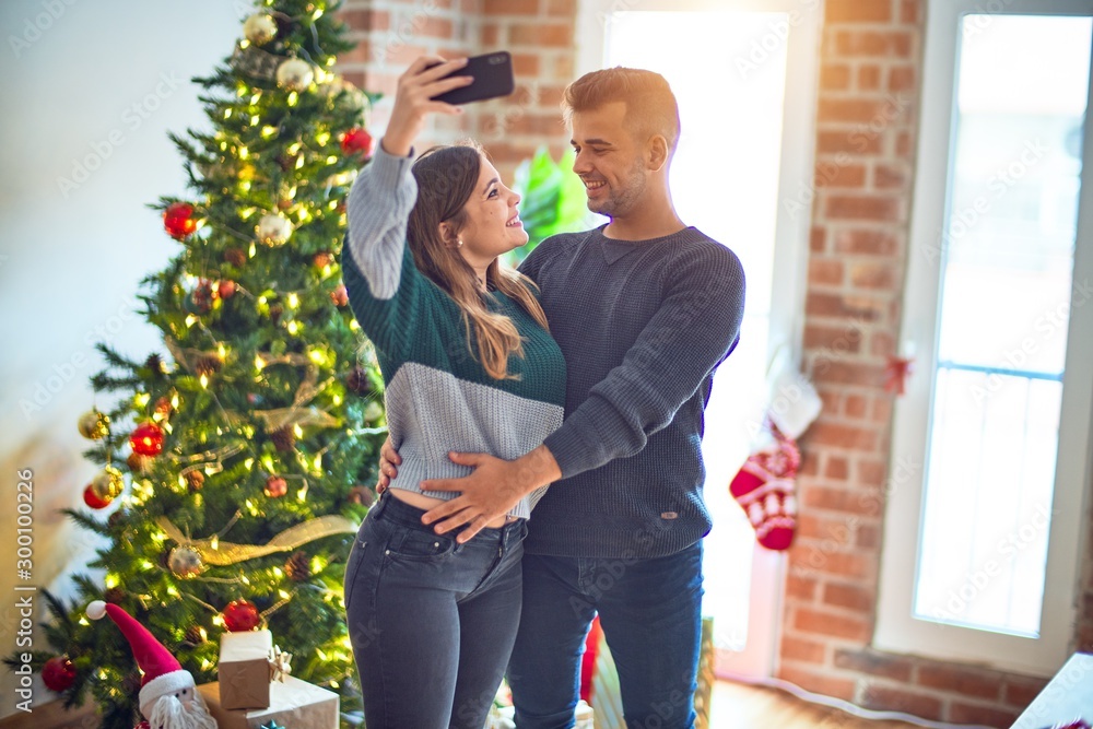 Young beautiful couple smiling happy and confident. Standing and hugging make selfie by camera around christmas tree at home Young beautiful couple smiling happy and confident. Standing and hugging make selfie by camera around christmas tree at home