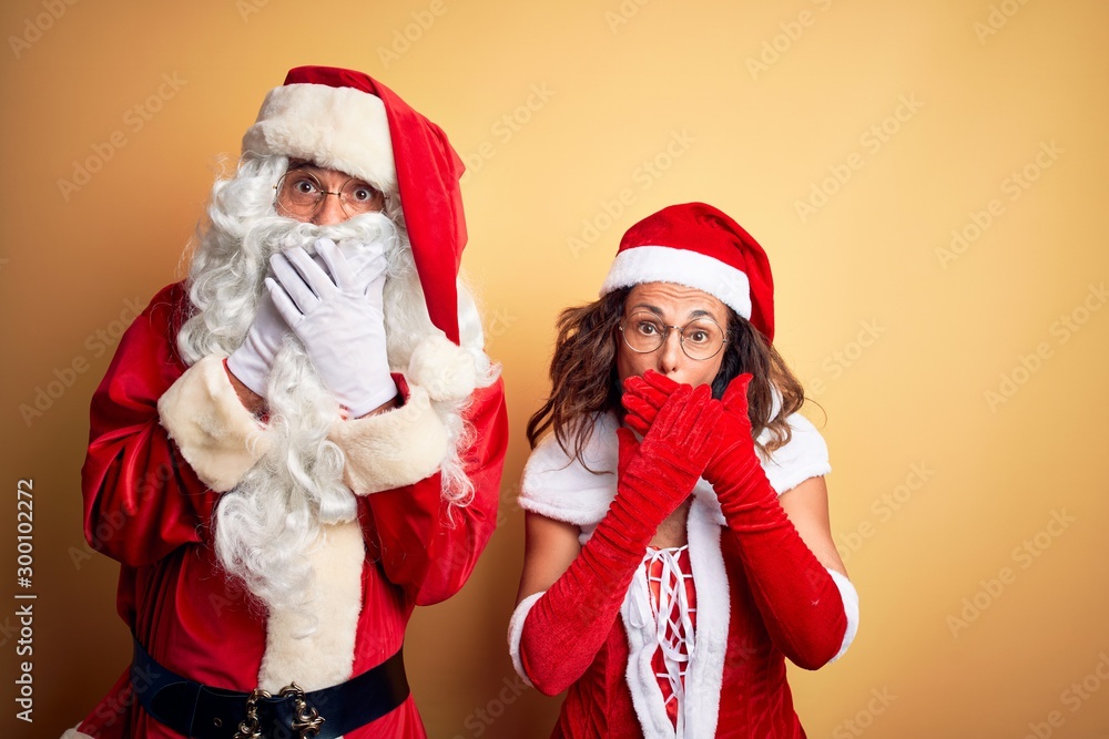 Fototapeta premium Middle age couple wearing Santa costume and glasses over isolated yellow background shocked covering mouth with hands for mistake. Secret concept.