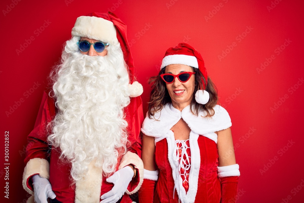 Middle age couple wearing Santa costume and sunglasses over isolated red background winking looking at the camera with sexy expression, cheerful and happy face. Middle age couple wearing Santa costume and sunglasses over isolated red background winking looking at the camera with sexy expression, cheerful and happy face.