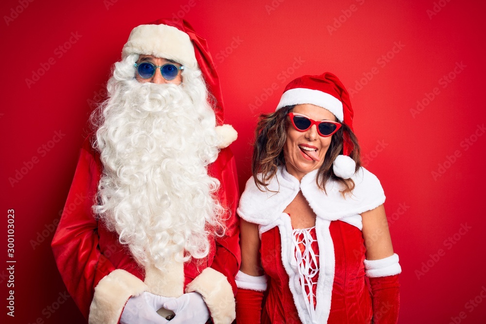 Middle age couple wearing Santa costume and sunglasses over isolated red background sticking tongue out happy with funny expression. Emotion concept. Middle age couple wearing Santa costume and sunglasses over isolated red background sticking tongue out happy with funny expression. Emotion concept.