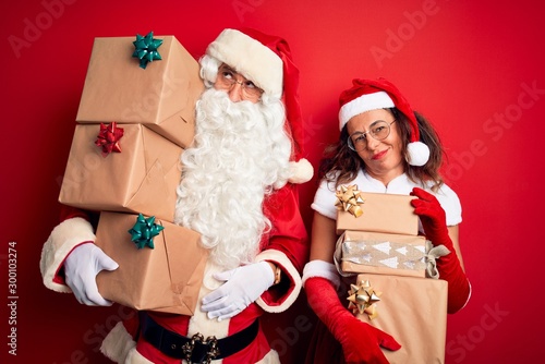 Middle age couple wearing Santa costume holding tower of gifts over isolated red background looking sleepy and tired, exhausted for fatigue and hangover, lazy eyes in the morning.