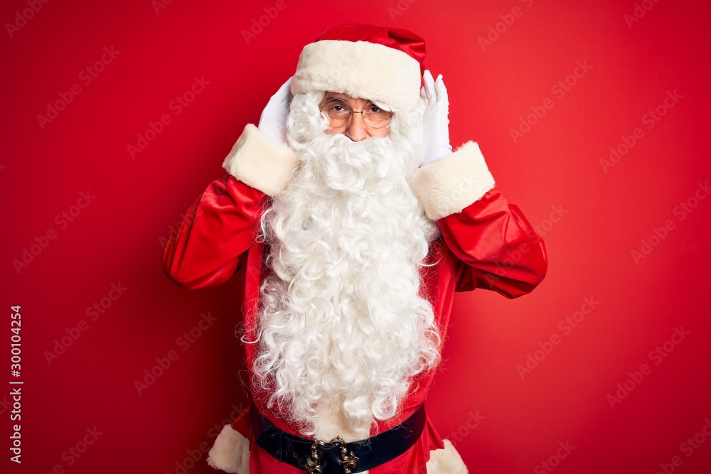 Middle age handsome man wearing Santa costume standing over isolated red background Trying to hear both hands on ear gesture, curious for gossip. Hearing problem, deaf