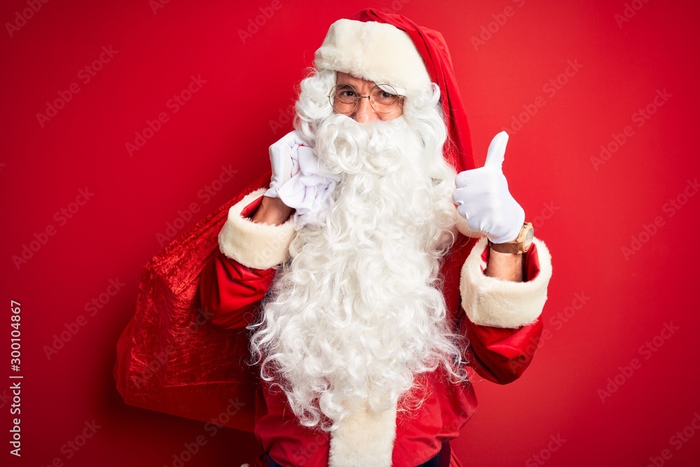 Middle age man wearing Santa costume holding sack with gifts over isolated red background happy with big smile doing ok sign, thumb up with fingers, excellent sign