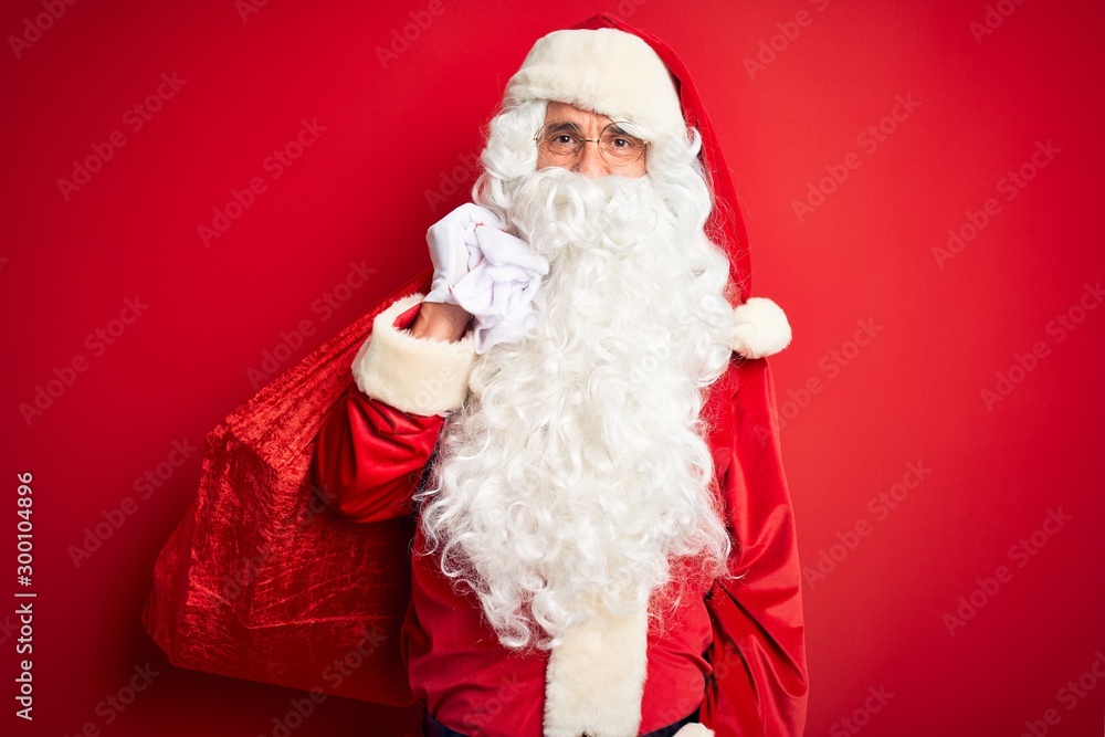 Middle age man wearing Santa costume holding sack with gifts over isolated red background with a confident expression on smart face thinking serious