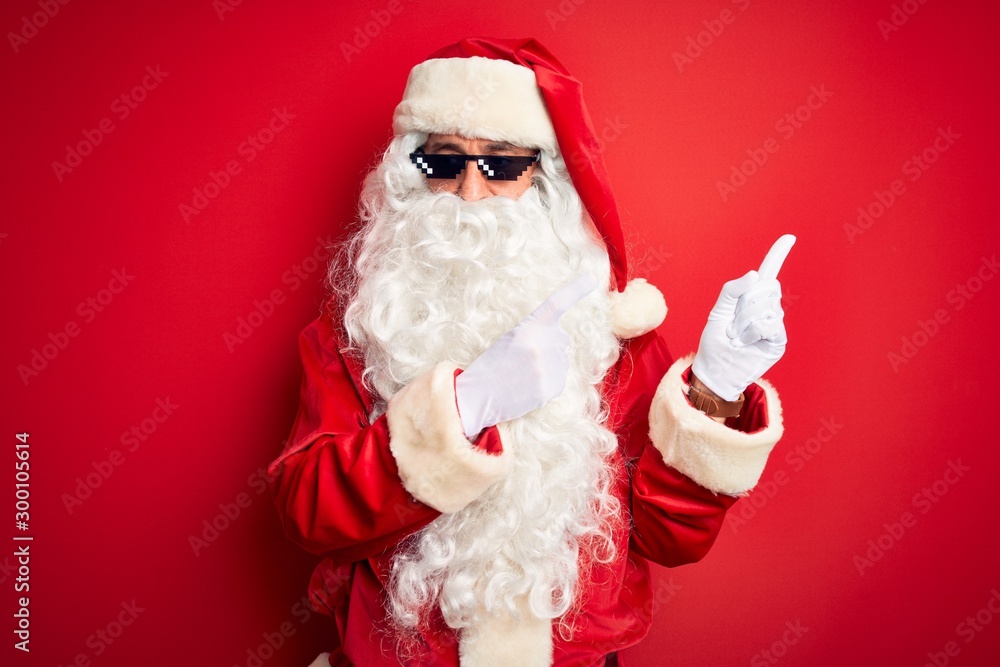 Middle age man wearing Santa Claus costume and sunglasses over isolated red background smiling and looking at the camera pointing with two hands and fingers to the side.