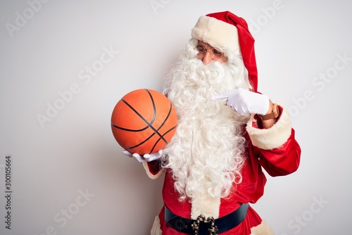 Senior man wearing Santa Claus costume holding basketball ball over isolated white background very happy pointing with hand and finger