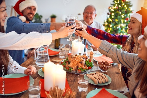 Beautiful family wearing santa claus hat meeting smiling happy and confident. Eating roasted turkey toasting with cup of wine celebrating Christmas at home