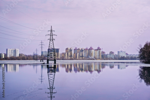 High-voltage power line over the wide lake and city in the distance. Sunset and reflection in water. Electrical conductor. Technology in the city. Voronezh  Russia.