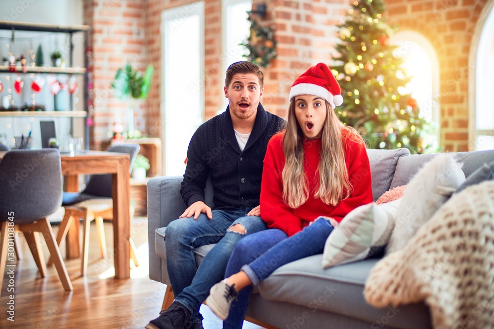 Young couple wearing santa claus hat sitting on the sofa around christmas tree at home afraid and shocked with surprise expression, fear and excited face.