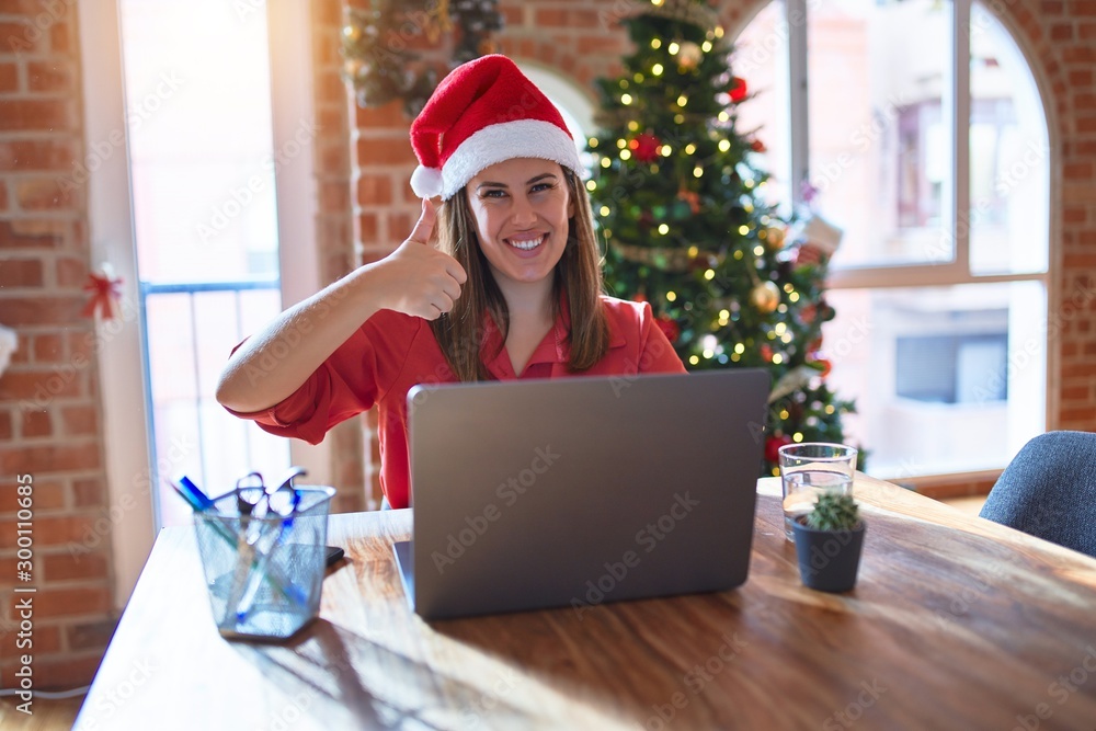 Beautiful woman sitting at the table working with laptop wearing santa claus hat at christmas doing happy thumbs up gesture with hand. Approving expression looking at the camera with showing success. Beautiful woman sitting at the table working with laptop wearing santa claus hat at christmas doing happy thumbs up gesture with hand. Approving expression looking at the camera with showing success.