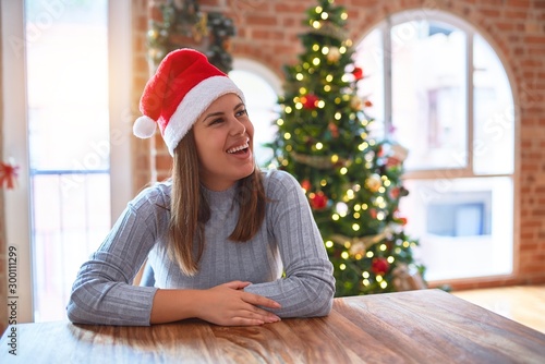 Young beautiful woman wearing santa claus hat at the table at home around christmas decoration looking away to side with smile on face, natural expression. Laughing confident.